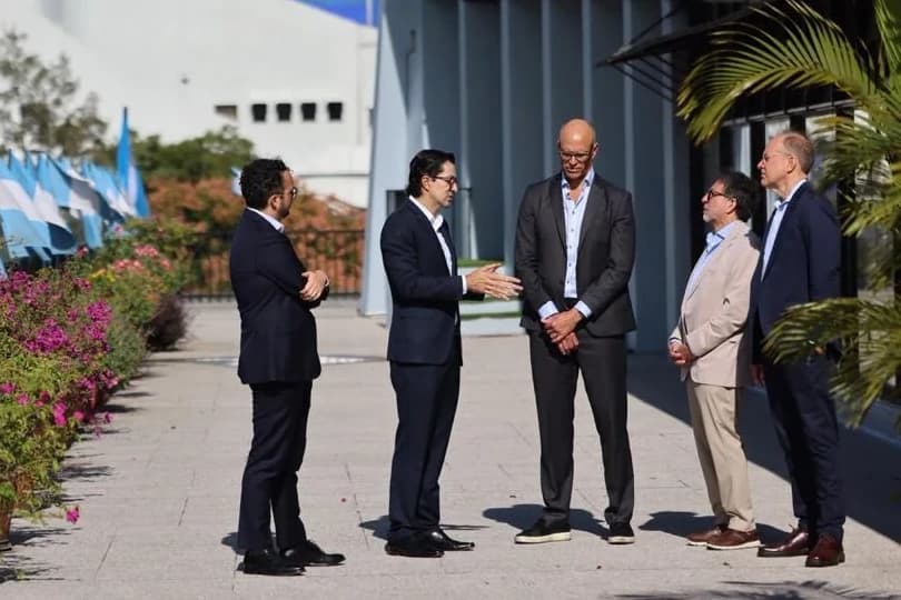 Cinco personas conversando de pie en una terraza soleada en la Ciudad de Guatemala, con una hilera de banderas guatemaltecas junto al muro